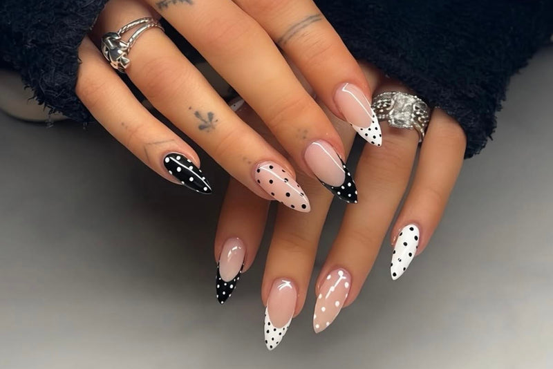 A close-up of hands with long stiletto-shaped nails featuring a mix of solid pale pink, white with black polka dots, and white with black French tips. The hands are adorned with multiple silver rings, and the person is wearing a dark fuzzy sleeve.